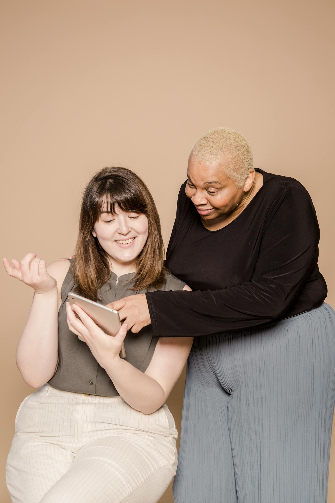 Positive plus size multiracial female coworkers surfing internet on modern tablet while standing on beige background in light studio during work together