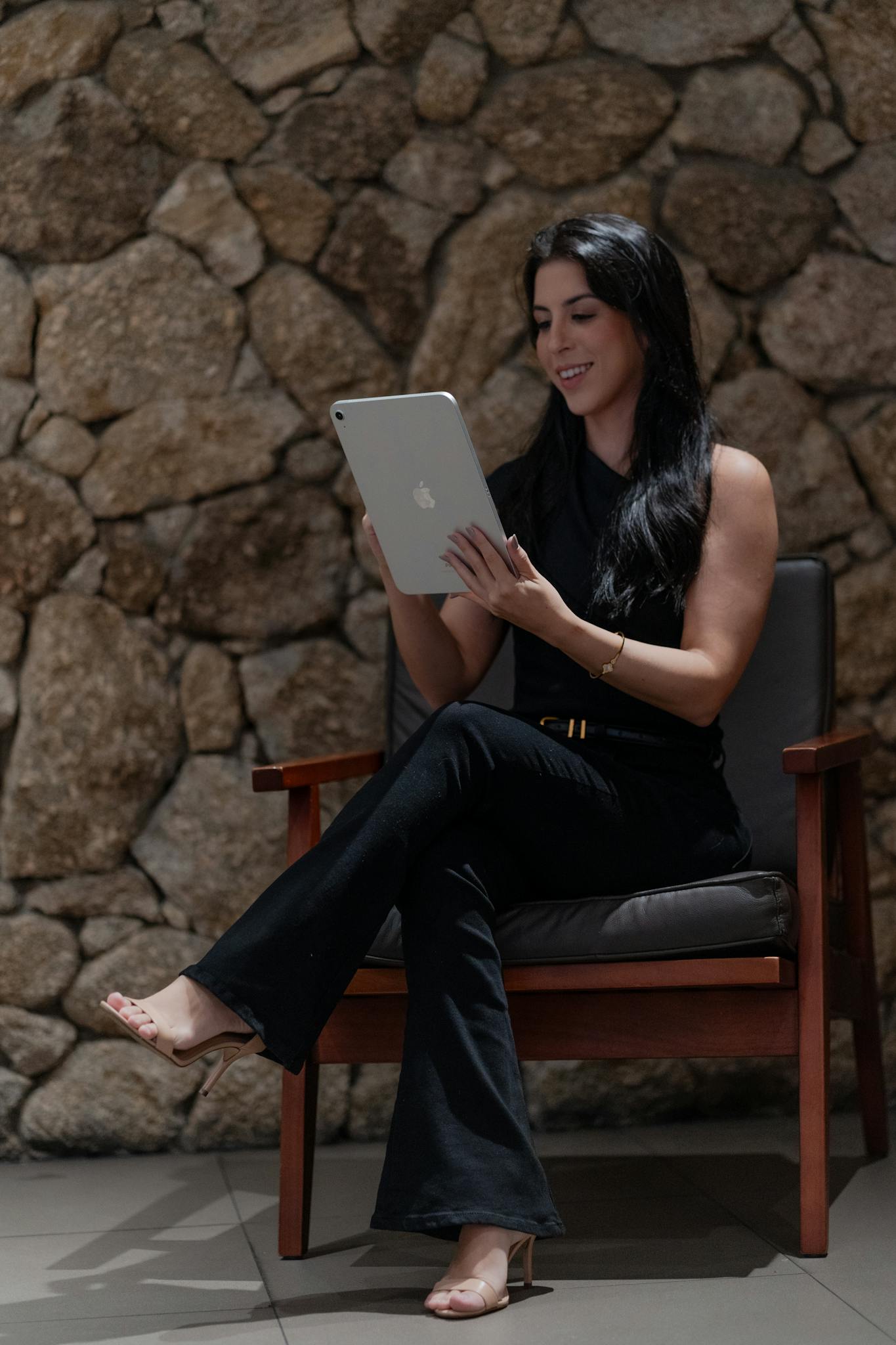 Smiling woman seated in a modern chair using a tablet against a stone wall backdrop.
