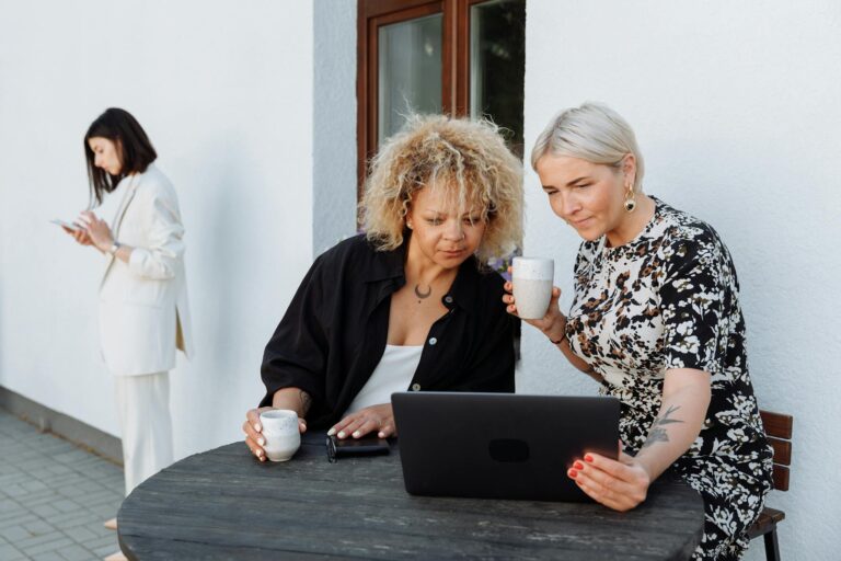 Women enjoying coffee while working on a laptop outside, teamwork in action.